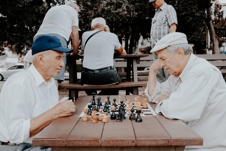 Two elderly men wearing caps play chess at an outdoor wooden table in a park, reminiscent of the lively social gatherings often found at Adult Day Centers, with other people seated and standing in the background.