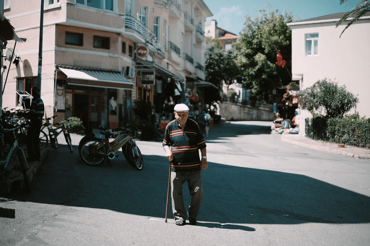 An elderly man wearing a cap and striped sweater walks with a cane across a sunny street in a quiet neighborhood with shops and bicycles—a peaceful scene where older people sometimes lose their way.