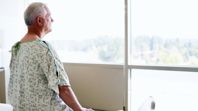 An older man in a hospital gown sits on a bed, looking out of a large window during the daytime, reflecting on his experience with inpatient services.