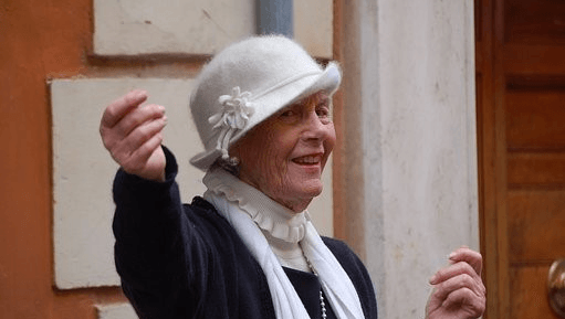 An older woman wearing a white hat and scarf raises one hand while smiling and standing outdoors near a building, showing how seniors maintain a positive outlook to enhance their quality of life.