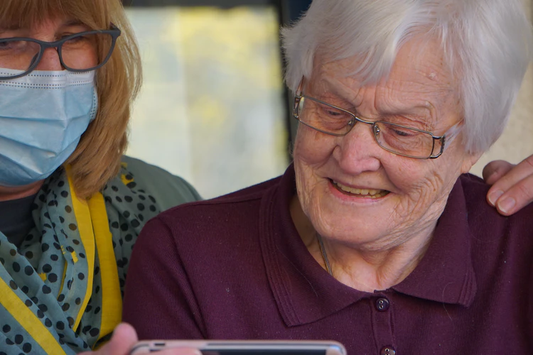 Two elderly women, one in a face mask and glasses, look at a smartphone together. The woman without a mask smiles—capturing a moment of connection during the Coronavirus Response on March 31, 2020.