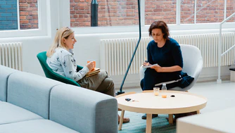 Two women sit in modern chairs at a round table, discussing questions to ask about an assisted living home in a bright, open room with brick walls and large windows.