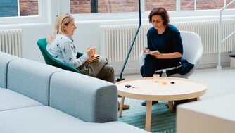 Two women, both experts, sit facing each other in a modern, bright room, engaged in conversation at a round wooden table with notebooks and cups, discussing ways to promote a high quality of life.