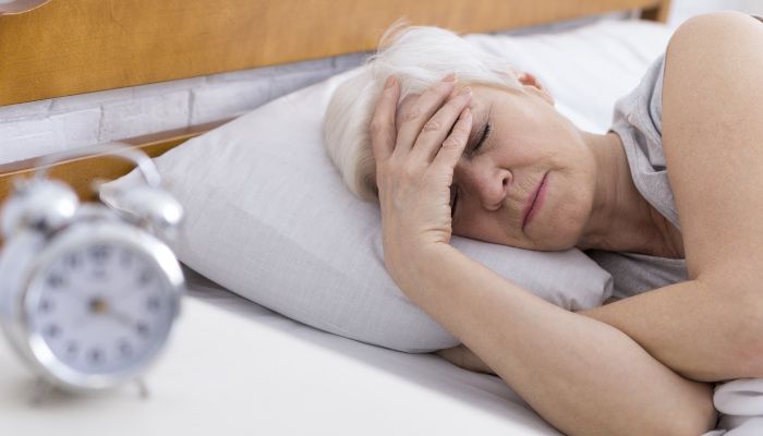 An older woman lies in bed with her hand on her forehead, appearing unwell or tired, possibly experiencing the effects of a disrupted sleep cycle, as an alarm clock rests on the bedside table in the foreground.