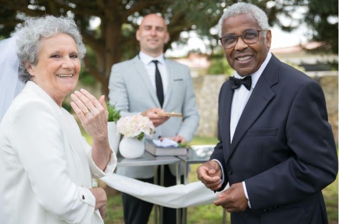 An older couple smiles at an outdoor wedding ceremony, receiving marriage advice from the officiant standing behind them holding a clipboard—a joyful moment for senior couples getting married.