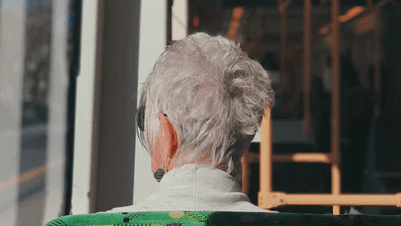 A person with short gray hair and earrings, possibly experiencing memory loss, sits on a green seat near a window inside a sunlit bus or tram, facing away from the camera.