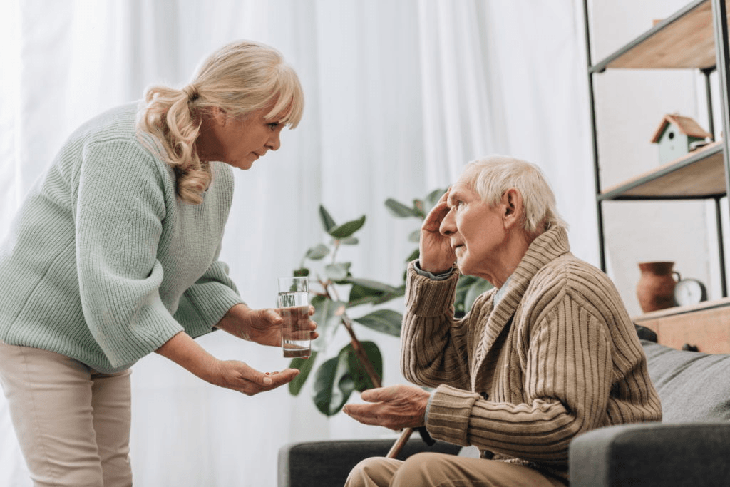 An older woman hands a glass of water to an older man sitting on a couch, touching his forehead—an everyday moment that helps in debunking Alzheimer’s myths by showing care and support in daily life. They are indoors near a plant and bookshelf.