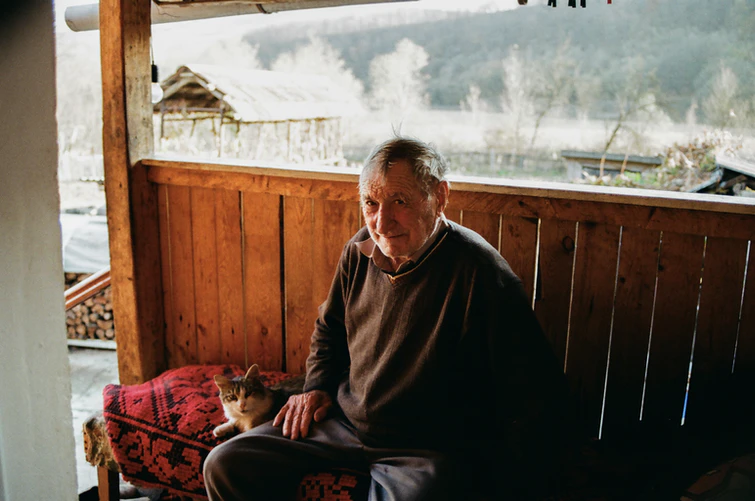An elderly man with dementia sits on a patterned bench beside a therapy animal cat on a porch with wooden railing, a rural landscape in the background.