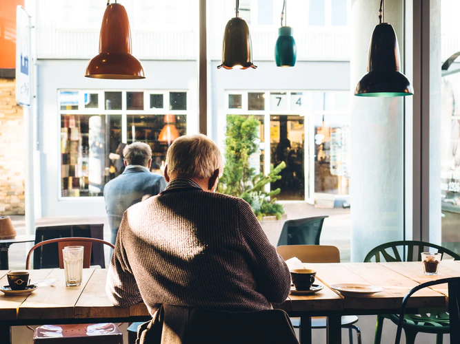 An older person, living with Alzheimer’s, sits alone at a café table with coffee and a glass of water, facing large windows that look out onto a sunny street.