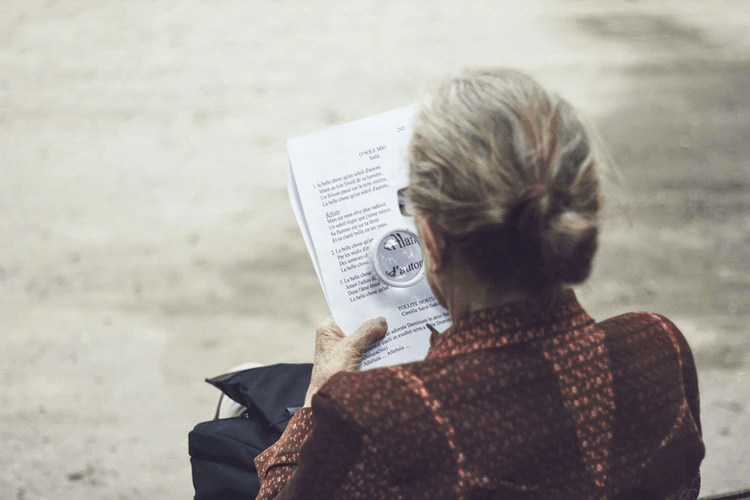 An elderly woman with gray hair in a bun, representing America's aging population, is reading a printed document outdoors while wearing a patterned brown top.