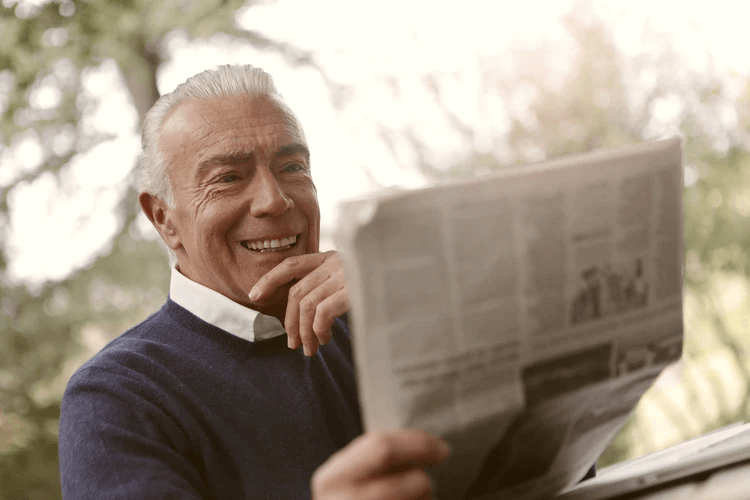 An older man in a blue sweater sits outside, smiling and holding a newspaper, enjoying the sunlight—a peaceful moment that can help reduce stress for both him and his caregiver.