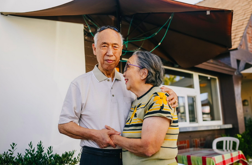 An older man and woman stand outside holding hands and smiling at each other, with a building and umbrella in the background—capturing the joy of a fresh start with senior-focused moving services.