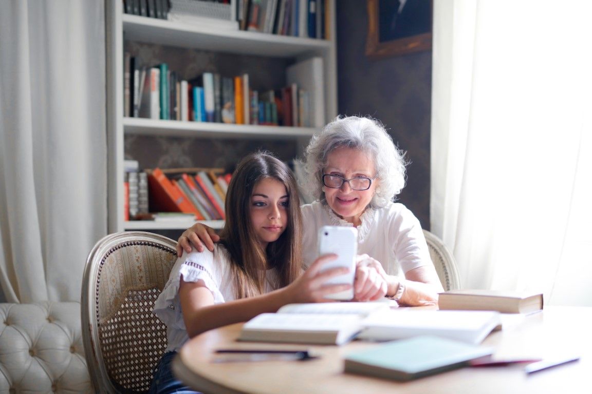 An older woman and a young girl sit together at a table, enjoying a virtual visit on a smartphone, with books and shelves in the background.