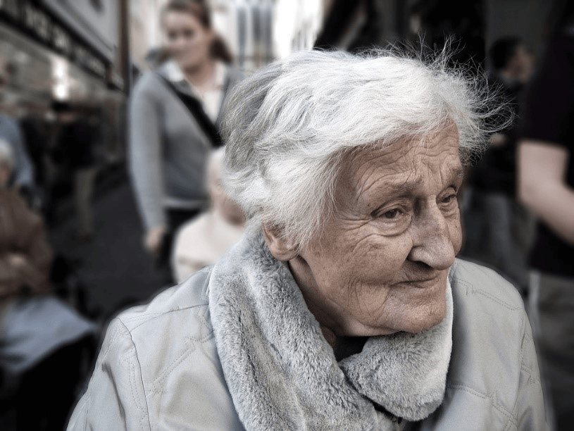 Elderly woman with white hair and a light gray coat stands outdoors, reflecting the quiet strength of those living with age-related memory loss; blurred people and buildings fill the background.