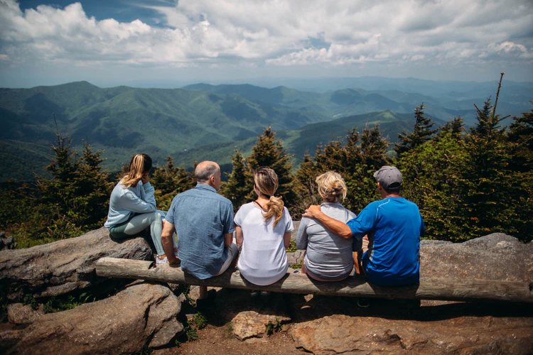 Five people sit on a log bench overlooking a scenic mountain landscape, enjoying the holidays together beneath green trees and a cloudy sky—creating memories that endure, even in the face of Alzheimer’s.