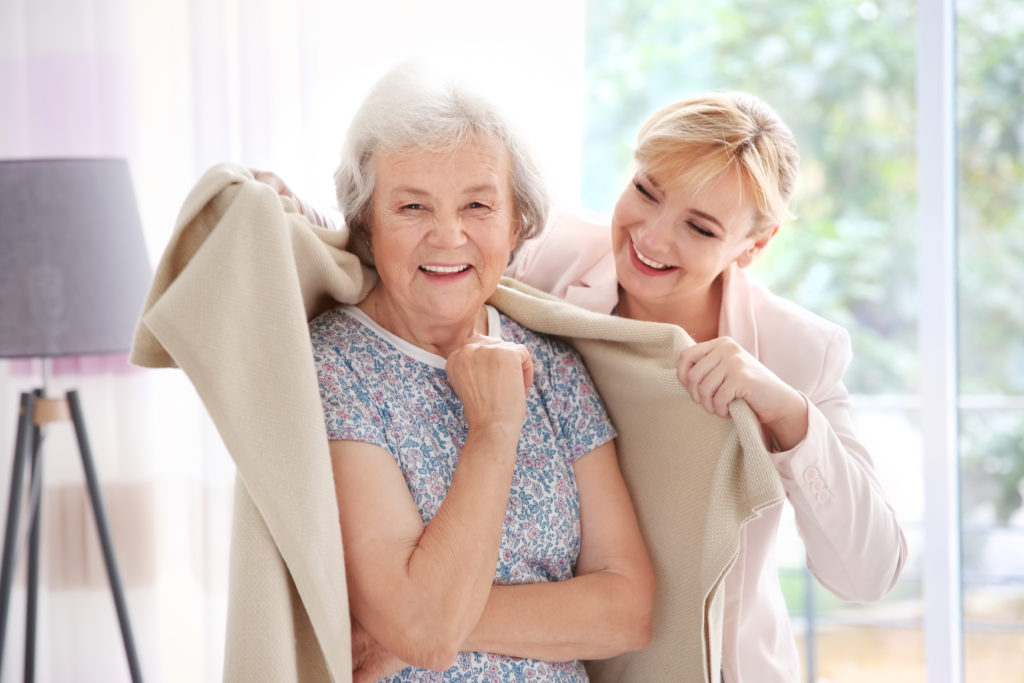 An older woman smiles as a younger woman drapes a beige blanket over her shoulders in a bright, indoor assisted living setting.