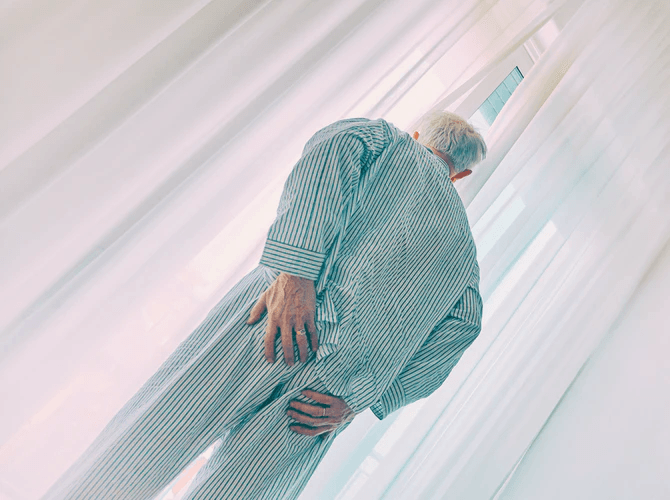 An older man in striped pajamas stands by a window with sheer curtains, holding his lower back with one hand—a scene often seen in individuals coping with Alzheimer’s disease.