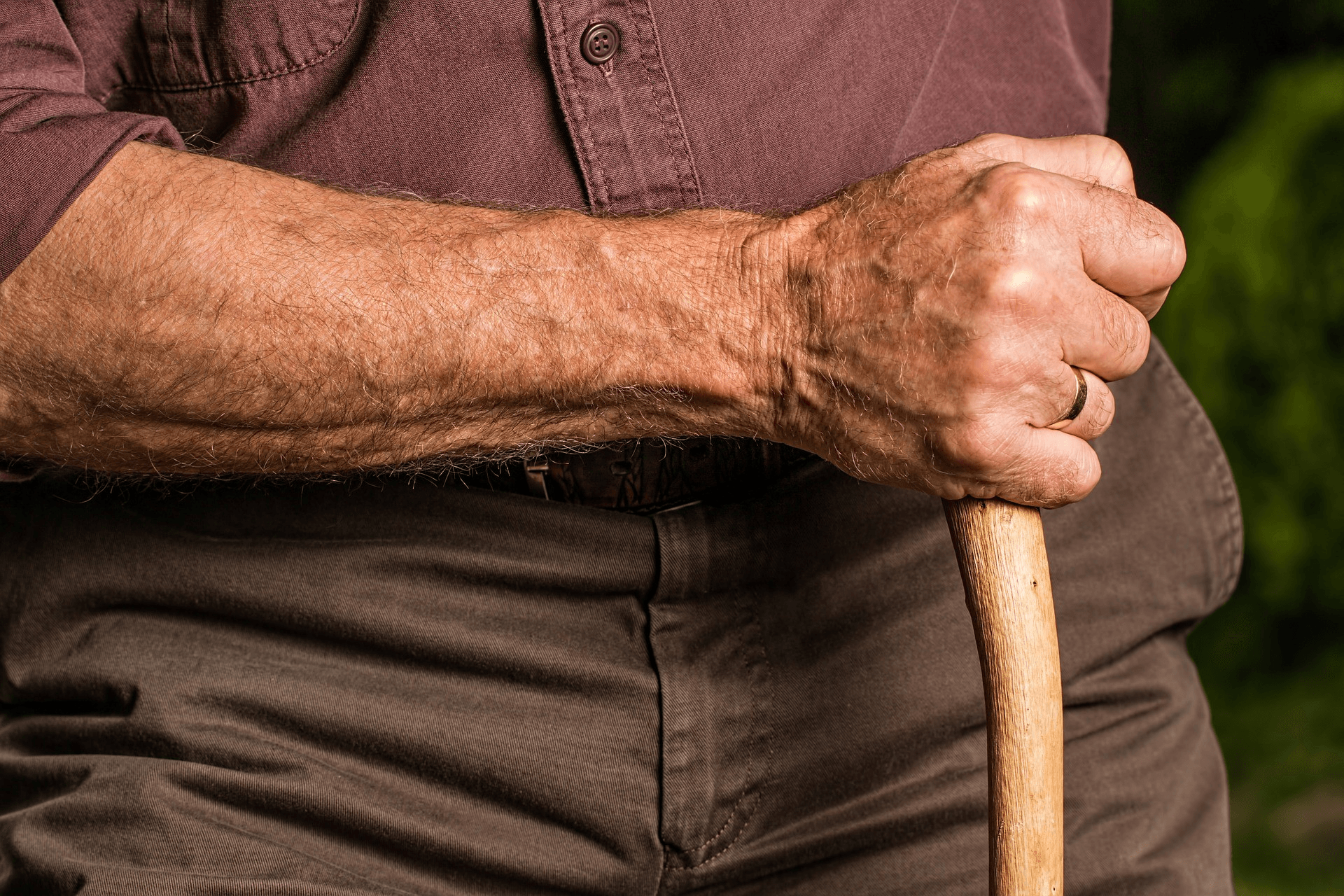 Close-up of an elderly person's hand gripping a wooden cane, wearing a brown shirt and pants—a quiet moment that reflects aging gracefully.