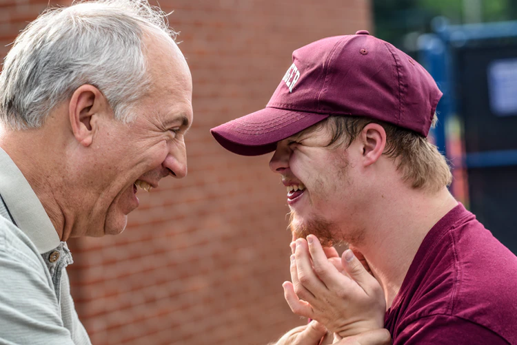 An older man and a young man wearing a maroon cap smile and laugh together outdoors near a brick wall, enjoying one of their favorite activities at home.