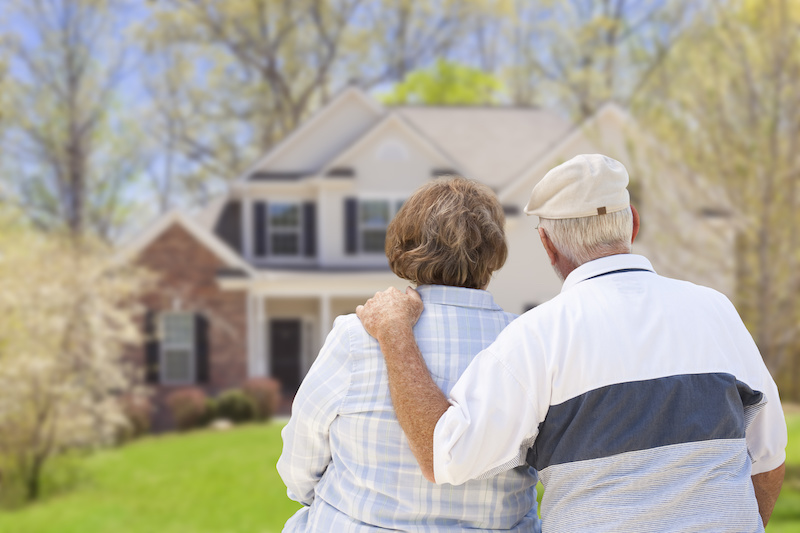 An elderly couple stands together outside, looking at a suburban house in the background surrounded by trees—reflecting on important home selling tips for seniors considering their next move.
