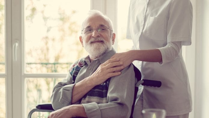 An elderly man with Alzheimer’s in a wheelchair is being assisted by a caregiver who rests a hand on his shoulder. They are indoors near a bright window, highlighting the importance of compassionate health care.