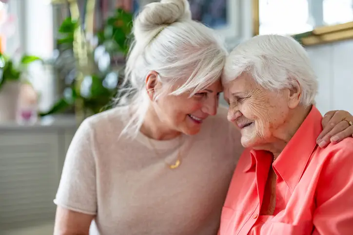 Two older women sit closely together, smiling and touching foreheads, in a warmly lit indoor setting with plants in the background. Two older women sit closely together, smiling and touching foreheads, in a warmly lit indoor setting with plants in the background.