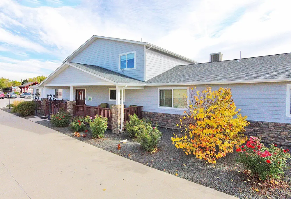 Arvada Blue House – 6741 W 65th Ave (ASL 11) A single-story building with light gray siding, stone accents, a front porch, and landscaped shrubs and trees, viewed from an angled perspective on a clear day.
