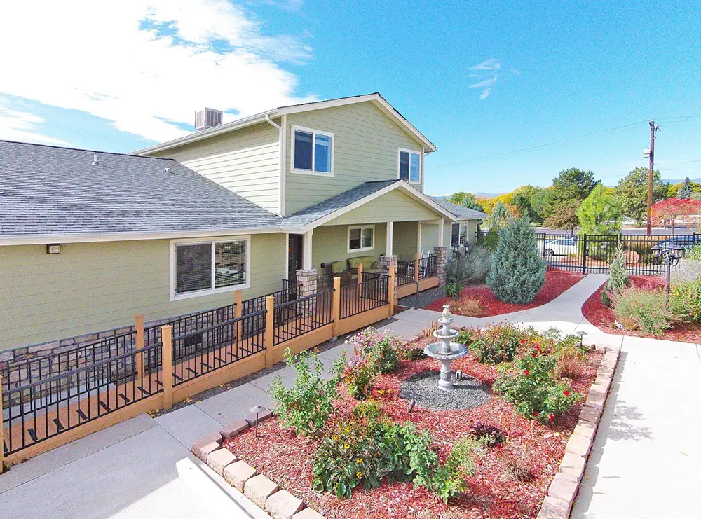 Arvada Green House – 6751 W 65th Ave (ASL 13) Two-story beige building with fenced front yard, landscaped garden, and a central water fountain under a clear blue sky.