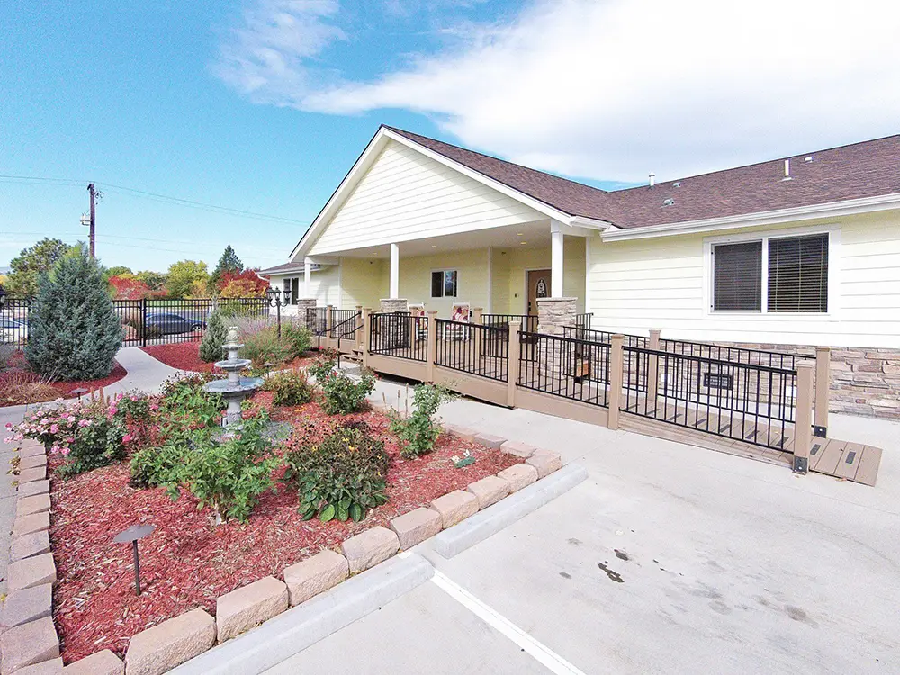 Arvada Peach House – 6761 W 65th Ave (ASL 12) Single-story building with a covered porch, wheelchair-accessible ramp, gated entry, and landscaped garden featuring a small fountain and flower beds.