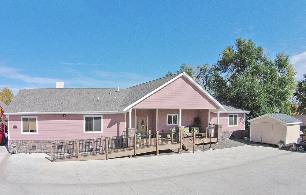 Arvada Rose House – 6735 W 65th Ave (ASL 15) Single-story pink house with front porch, ramp, gabled roof, and a small storage shed in the background, set against a clear blue sky.