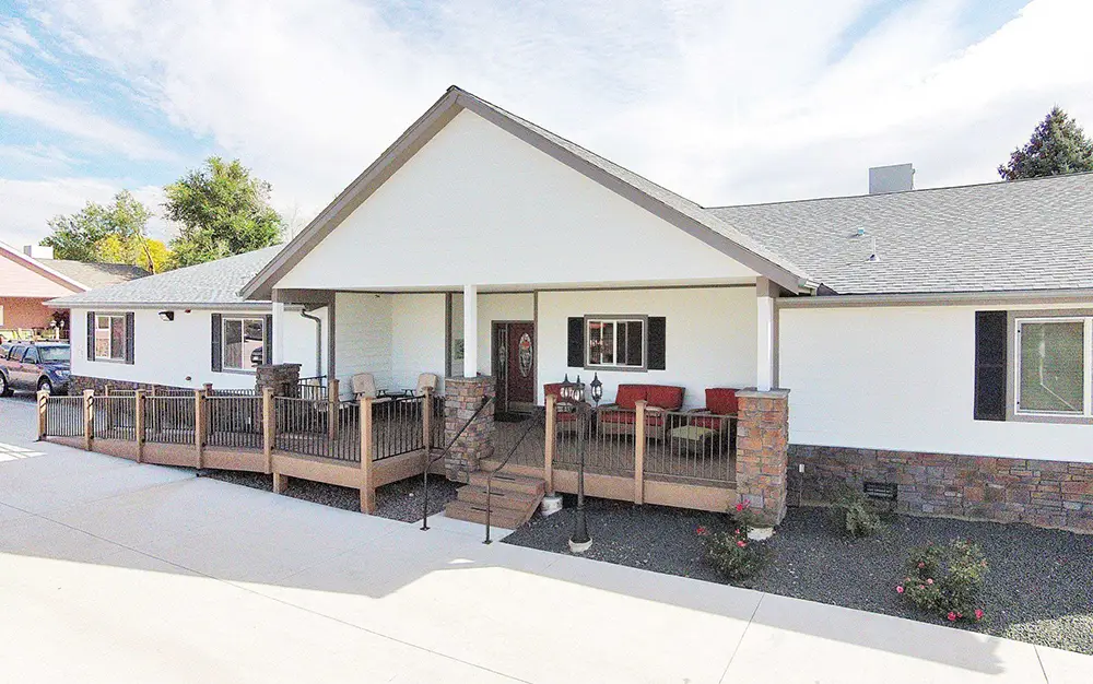 Arvada White House – 6725 W 65th Ave (ASL 16) Single-story white house with a gable roof, front porch with ramp and stairs, stone columns, and outdoor seating area.