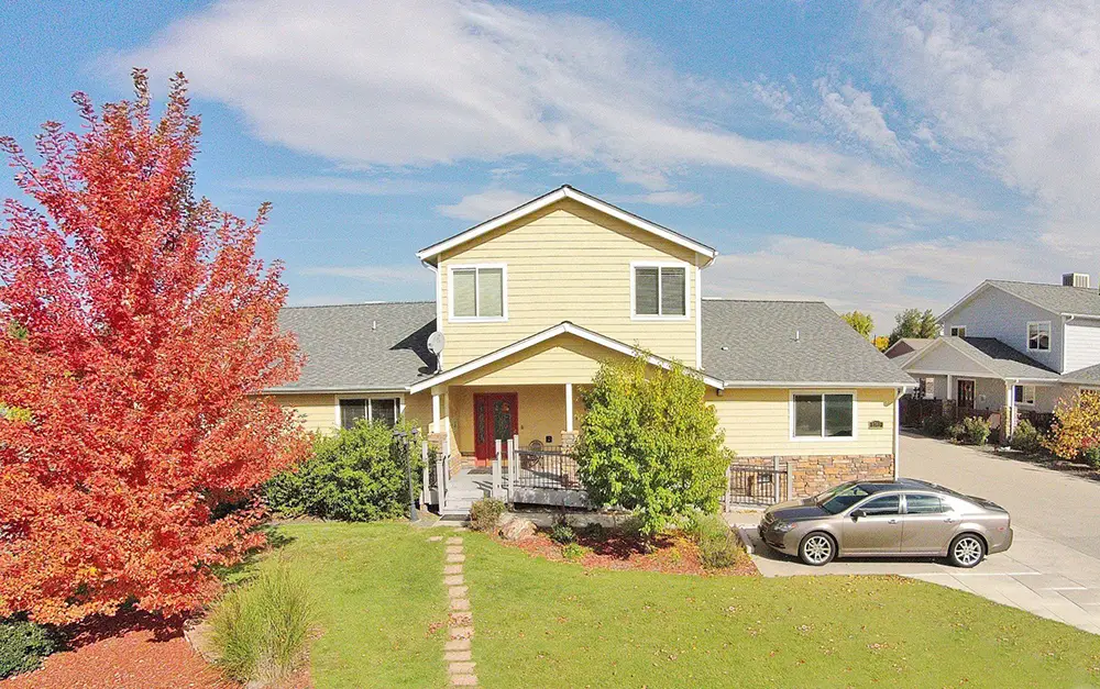 Arvada Yellow House – 6745 W 65th Ave (ASL 14) A two-story yellow house with a gray roof, front porch, and red front door, has a silver car parked in the driveway and a red-leafed tree in the yard under a blue sky.