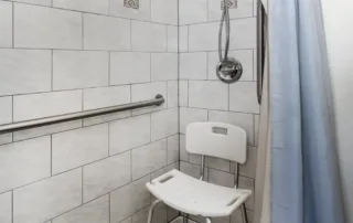 Arvada-gallery-1 Accessible shower with a white tiled wall, silver grab bar, handheld showerhead, light blue shower curtain, and a white shower chair.