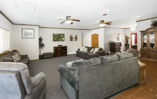 Arvada-gallery-10 Spacious living room with gray sofas, armchairs, a wooden cabinet, ceiling fans, wall art, and a carpeted floor. Natural light enters through windows on the left.