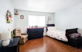 Arvada-gallery-12 A tidy bedroom with wood flooring, a single bed with floral bedding, a cushioned chair, a black dresser, a side table with a lamp, and a window letting in natural light.