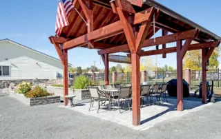 Arvada-gallery-18 A wooden pergola with an American flag covers a patio area with two outdoor dining tables and chairs, surrounded by a landscaped yard and a fence.