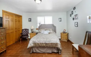 Arvada-gallery-2 A bedroom with a double bed, wooden floor, bedside tables, leather chair, dresser, and framed photos on white walls. A window with blinds is behind the bed.