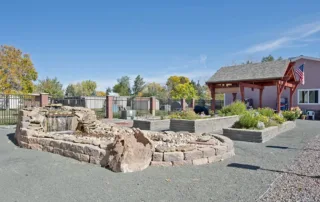 Arvada-gallery-20 Outdoor garden area with raised garden beds, a rock water feature, gravel pathways, and a wooden pergola beside a pink house under a clear blue sky.