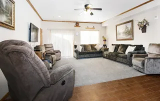 Arvada-gallery-3 A spacious living room with two gray sofas, two brown recliners, a wall-mounted TV, and framed artwork, featuring carpeted and tiled flooring.
