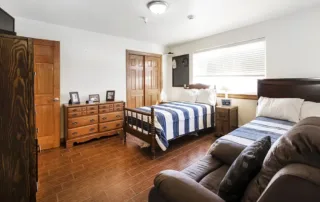 Arvada-gallery-5 Bedroom with two wooden twin beds, a brown recliner, wooden dresser and wardrobe, and a window with blinds letting in natural light.