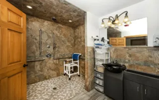 Arvada-gallery-7 Accessible bathroom with a roll-in shower, grab bars, a shower chair, a large mirror, sink, and storage shelves under warm overhead lighting.
