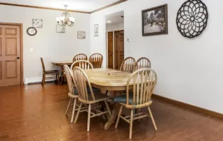 Arvada-gallery-9 Dining room with a wooden table and eight chairs, brown tile flooring, wall art, a clock, and a hanging light fixture. Another smaller table is visible in the background.