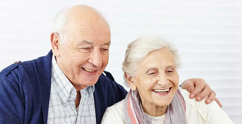 An elderly man and woman sit close together, smiling with the man's arm around her shoulder, in front of a white background—capturing the warmth and companionship found beyond just the cost of assisted living.