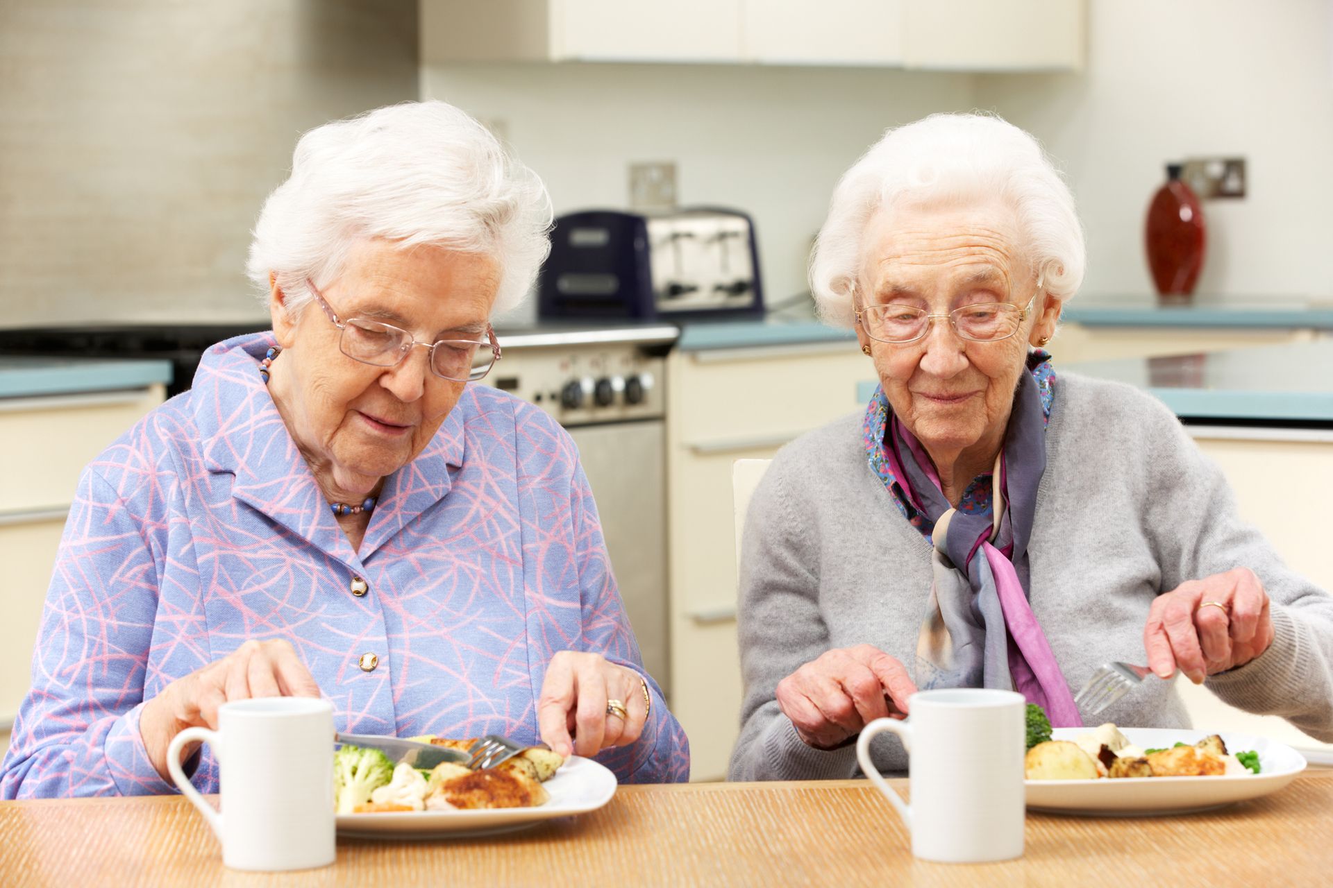 Two elderly women sit at a kitchen table eating a meal together, discussing senior living questions over plates of food and mugs in front of them.