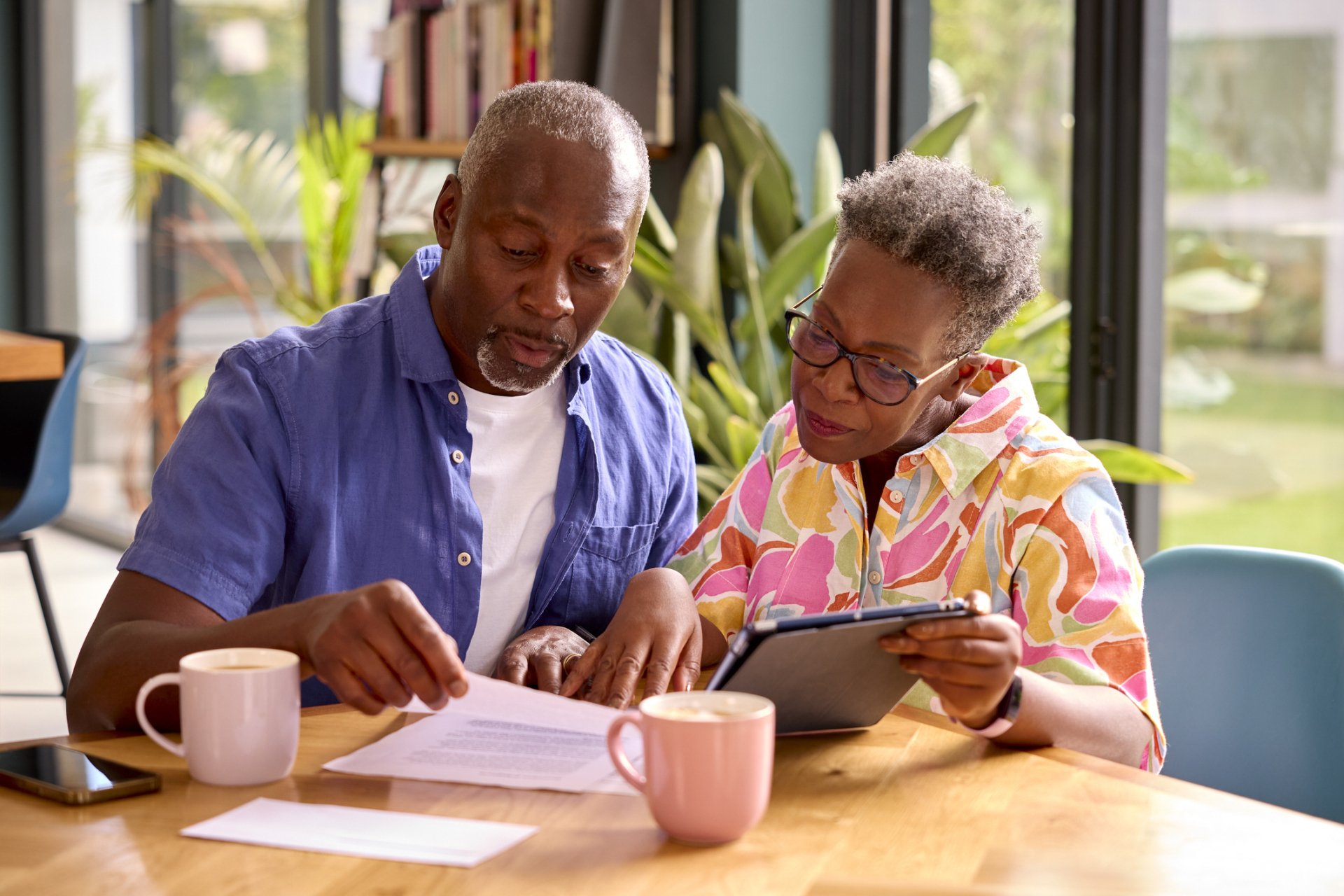 An older couple sits at a table with coffee cups, reviewing a document and using a tablet together in a bright indoor setting, exploring senior living options and home cost comparison.