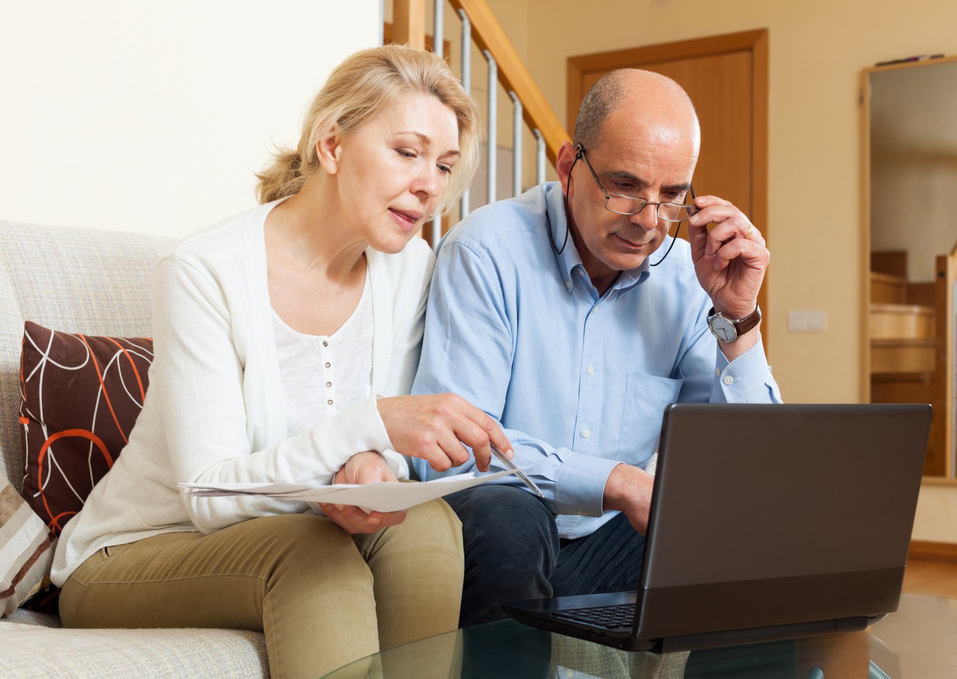 An older couple sits on a couch, reviewing papers and looking at a laptop together, appearing focused and engaged as they discuss funding senior living and managing senior living expenses.
