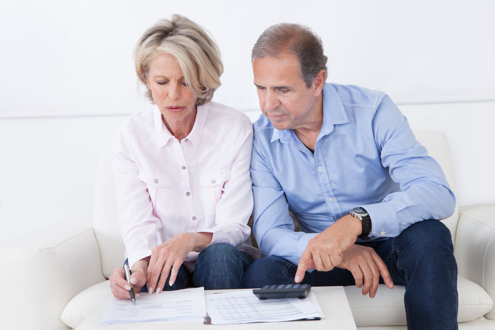 An older woman and man sit on a couch reviewing documents together, perhaps discussing senior care pricing; the woman writes on paper while the man uses a calculator.