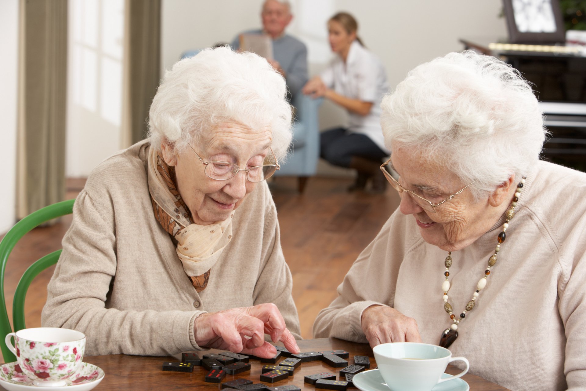 Two elderly women sit at a table in one of the memory care communities, playing dominoes with teacups in front of them; two more people are seated in the background.