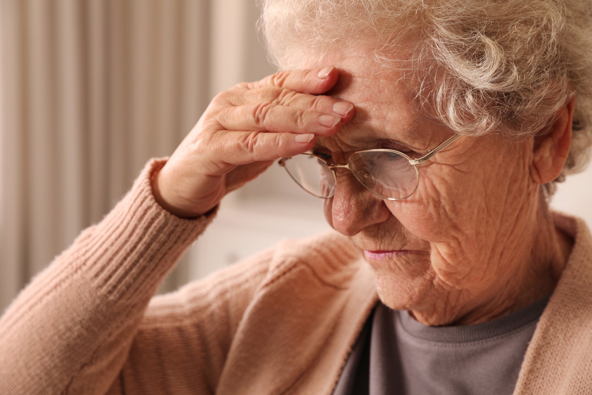 Elderly woman wearing glasses and a beige sweater holds her hand to her forehead, appearing concerned or thoughtful—possibly showing early signs that may prompt consideration of senior living for your loved one.
