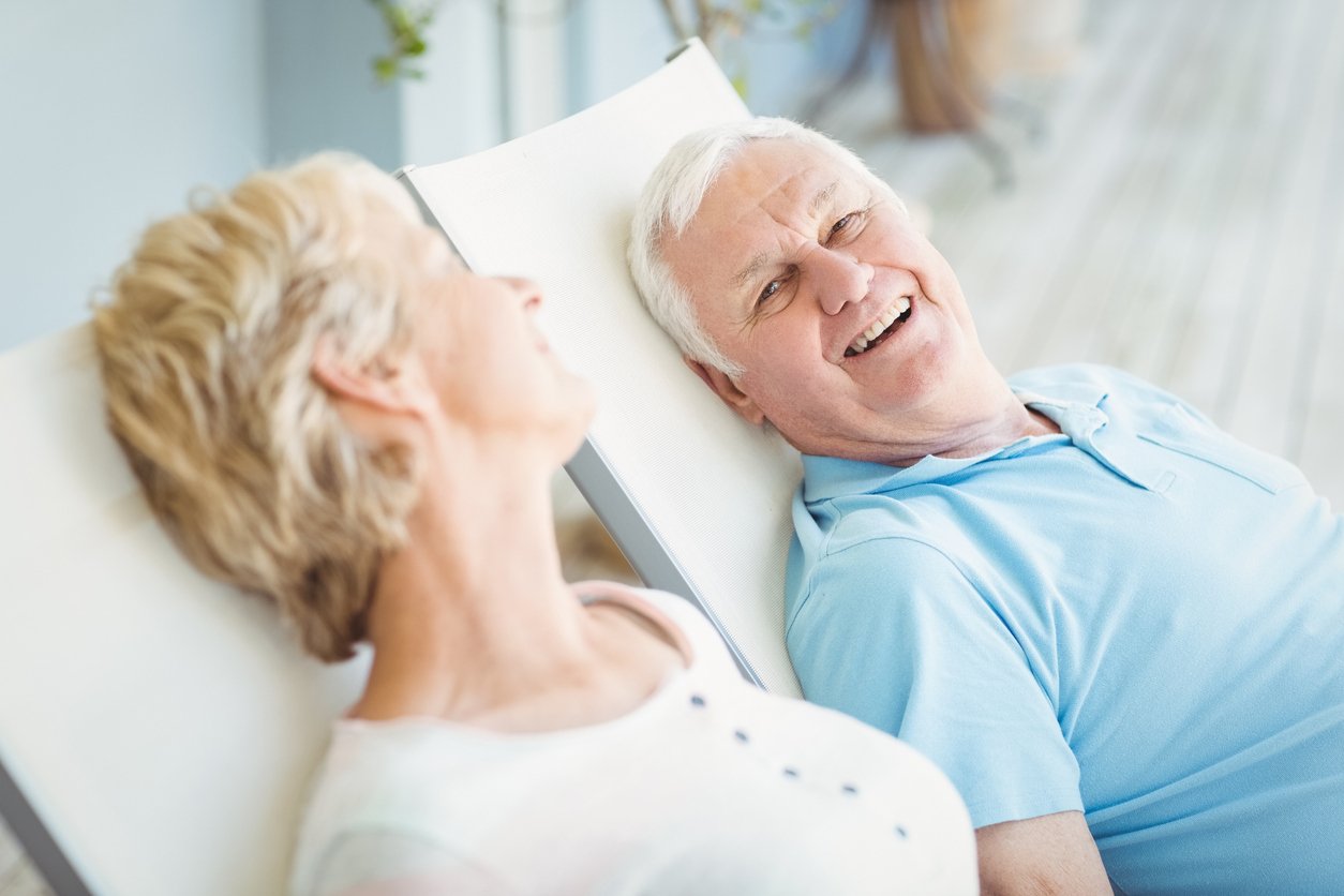 An older man and woman sit on outdoor lounge chairs, smiling and laughing together in a relaxed setting that highlights the comfort and amenities of senior living.